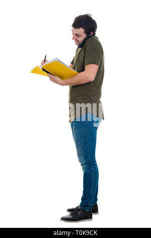 Full length of busy casual young man doing two things at a time, talking to phone and making notes in his copybook. Concerned guy holding phone by a s Stock Photo