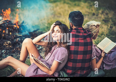 Camping in wilderness. Turn back guy looking at fire while two beautiful girls read book. Smiling brunette with braces leaning on her friend Stock Photo