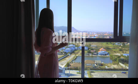 woman in a pink transparent dressing gown stands and looks out into the panoramic window and drinks coffee. 4k Hua Hin, Thailand. Stock Photo