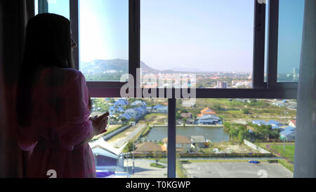 woman in a pink transparent dressing gown stands and looks out into the panoramic window and drinks coffee. 4k Hua Hin, Thailand. Stock Photo