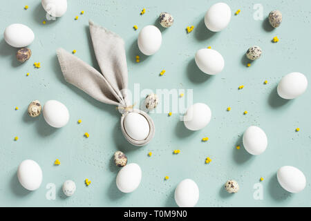 Pattern of chicken and quail eggs with bunny ears of linen napkin, small yellow flowers on the blue textured table, Easter composition, top view Stock Photo