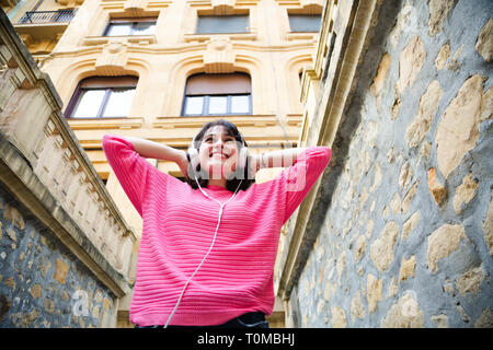 Happy young woman in pink sweater listening to music in the street. Low angle view Stock Photo