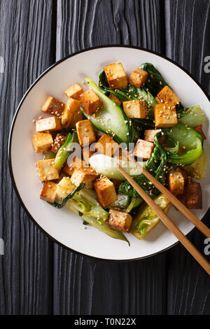 Traditional fried tofu with bok choy, soy sauce and sesame close-up on a plate on the table. Vertical top view from above