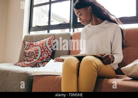 African girl sitting on a sofa with a tablet pc reading a book. Young student studying indoors at college campus. Stock Photo