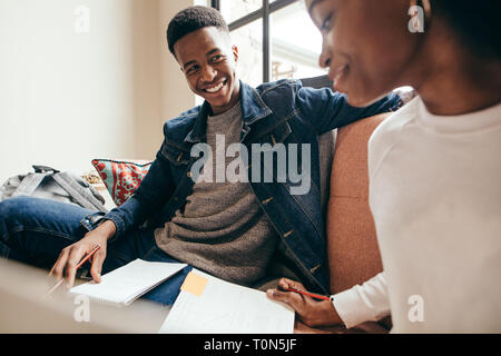 Smiling teenager looking at girl reading book on sofa at university campus. College students studying together in campus building. Stock Photo