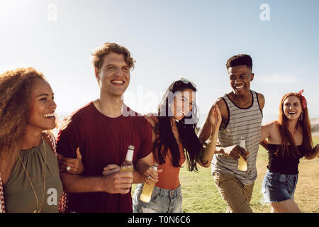 Multiracial group of young people having a great time together outdoor on a summer day. Young men and women with beers walking together. Stock Photo