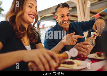 Smiling man and woman eating snacks sitting at a restaurant. Happy couple pouring tomato sauce on hot dogs sitting at a dining table. Stock Photo