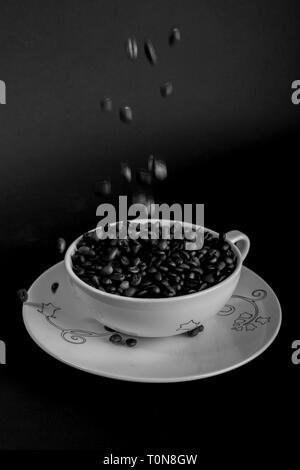 concept showing coffee beans being poured into a white coffee cup and saucer on black background Stock Photo