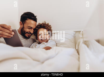 Man lying on bed with his son and talking. Cheerful father and son spending time together using a tablet pc. Stock Photo