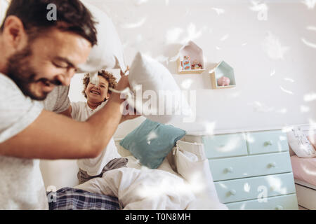 Cheerful man pillow fighting with son sitting on bed at home. Father and son having fun playing with pillows with feathers flying around. Stock Photo