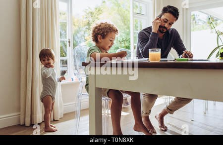 Man sitting at a table and talking over mobile phone with kids engaged in activity. Man working from home and babysitting kids. Stock Photo