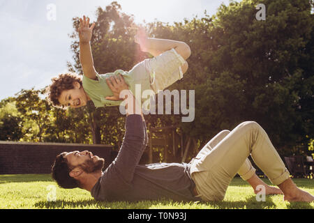 Man lying on ground and lifting his son with his arms in a park. Father and son playing in park on a sunny day. Stock Photo