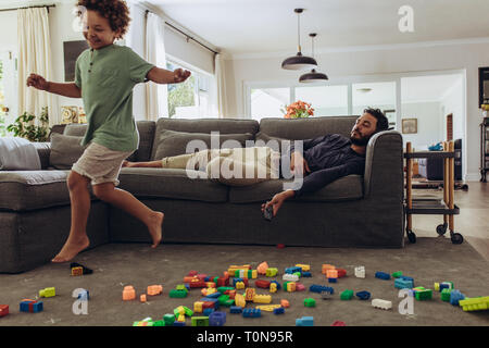 Man sleeping on couch while his kid is playing at home. Boys running around at home with building blocks spread on the floor. Stock Photo