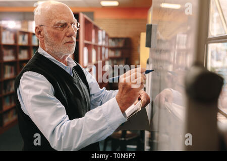 Side view of a university professor writing teaching in class. Senior lecturer writing on whiteboard in classroom. Stock Photo