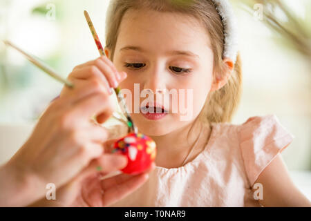 A mother and daughter celebrating Easter, painting eggs with brush. Cute little girl in bunny ears preparing the holiday. Stock Photo