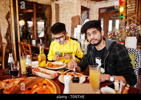 Asian friends guys eating pizza during party at pizzeria. Happy indian people having fun together, eating italian food and sitting on couch. Stock Photo