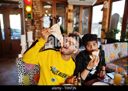 Asian friends guys eating pizza during party at pizzeria. Happy indian people having fun together, eating italian food and sitting on couch. Stock Photo