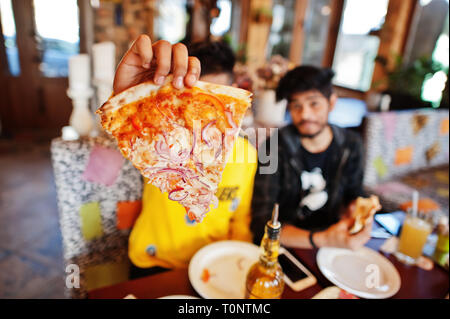Asian friends guys eating pizza during party at pizzeria. Happy indian people having fun together, eating italian food and sitting on couch. Stock Photo