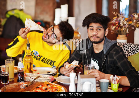 Asian friends guys eating pizza during party at pizzeria. Happy indian people having fun together, eating italian food and sitting on couch. Stock Photo