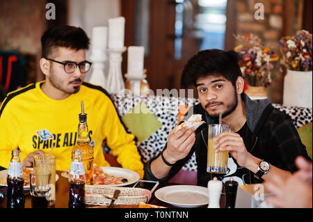 Asian friends guys eating pizza during party at pizzeria. Happy indian people having fun together, eating italian food and sitting on couch. Stock Photo