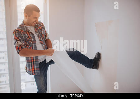 Funny man changes the house wallpaper. The worker or builder holds a cup of coffee in his hands and looks at the tablet. Against the background of con Stock Photo