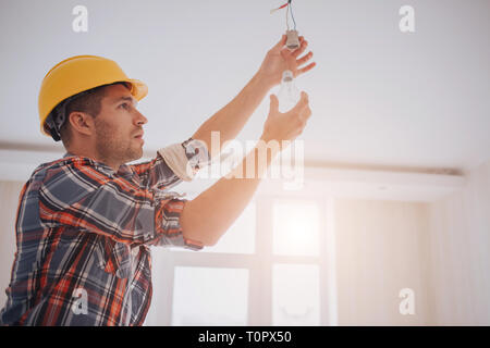 Handsome young builder in a yellow construction helmet is twisting the light bulb in. The man is looking up Stock Photo