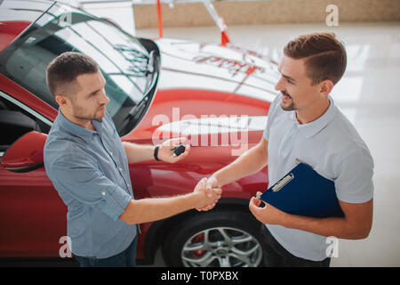 Serious men stand and shake each other's hands. Purchaser holds car key. Dealler hold tablet. They made a deal. Red car belongs to men on left Stock Photo