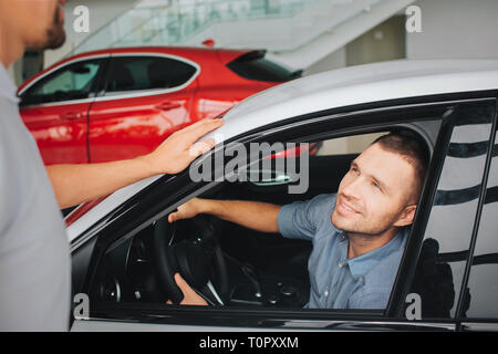 Handsome and satisfied young men sitting in white car and look at manager. He smiles. Guy hold hands on steering wheel. Bearded manager stands beside  Stock Photo