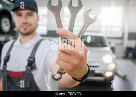 Young handsome man shows set of wrenches. He looks at them and smile. Guy wears uniform. He stands in front of white car. Young man smiles a bit Stock Photo