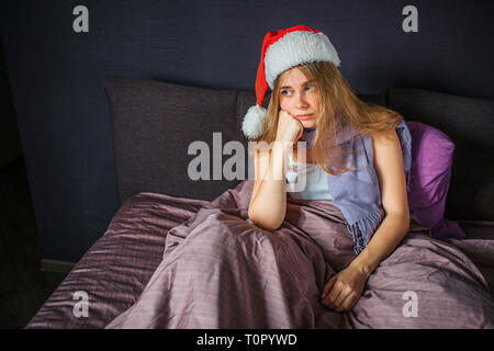 Sick young woman sits on bed and looks straight. She is bored. Woman holds hands under chin. She has Christmas hat on head. Stock Photo