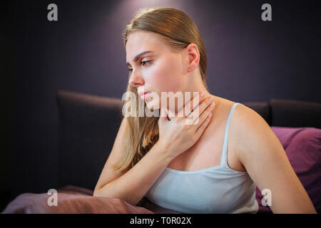 Worried young woman sits on bed alone and looks down. She holds hand on neck. Woman feels pain there. Stock Photo