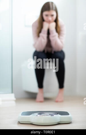 Unhappy Teenage Girl Sitting In Bathroom Looking At Scales Stock Photo