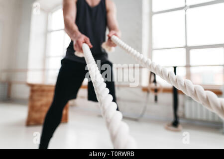 Muscular male athlete doing exercises with rope at gym. Fitness man pulling rope light hall Stock Photo
