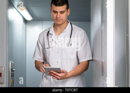Bright photo of male doctor in uniform with stethoscope coming out of the elevator and using computer tablet in hospital. Modern medical concept Stock Photo