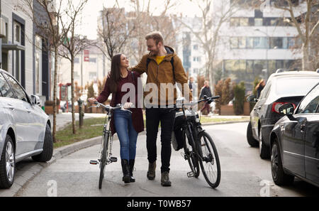 two friends or couple, 20-29 years, walking and talking on a street, pushing their bicycles, looking at each other. Stock Photo