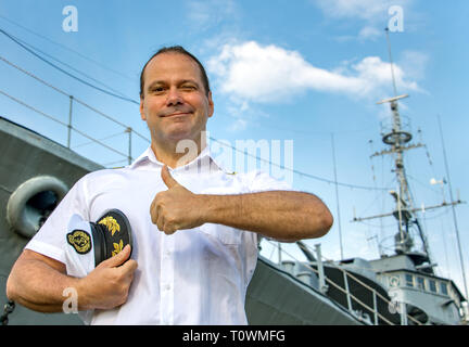 Captain standing in dock before warship and  shows thumb up. A sailor officer in white uniform stands beside battleship. Stock Photo