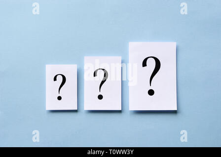 Three question marks of different size printed on white cards arranged in ascending order centered on a blue background in a conceptual image Stock Photo