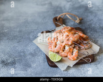 Red shrimps on metal skewers with lime juice on wood board on grey background. Stock Photo