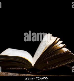 Old book open on vintage table on dark background. Read and study vintage bible with illuminated page. Literature and education still life. Stock Photo