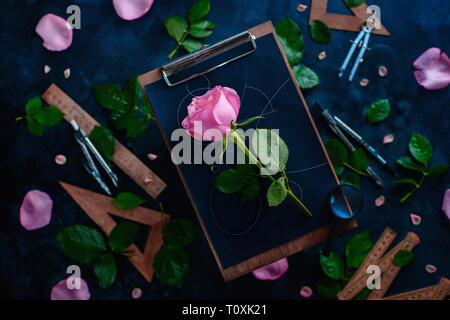 Rose flower with blueprint lines on a wooden clipboard with pencils, rulers, and compasses around. Nature and geometry conceptual flat lay Stock Photo