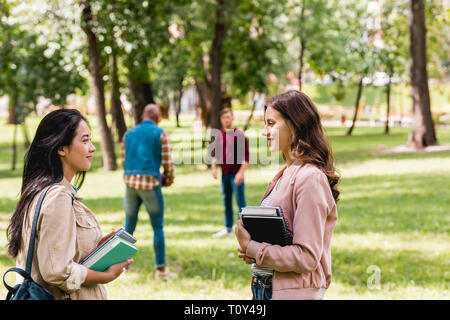 selective focus of cheerful girls talking while standing with books near students playing american football