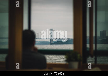 The George Washington Bridge crosses the Hudson River connecting Manhattan to New Jersey as seen through a window on a boat with a tourist enjoying the view on a hazy early autumn day. Stock Photo