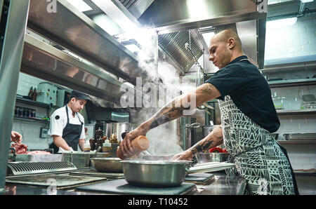 How to cook a meat Concentrated young chef in apron and cooks preparing food together in a restaurant kitchen. Cooking process Stock Photo