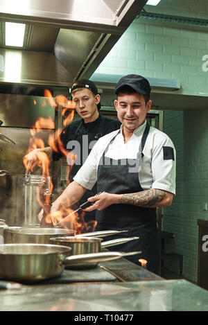 Working with fire. Two young and positive cooks learning how to make a flambe on food in restaurant kitchen. Culinary school. Teamwork. Stock Photo