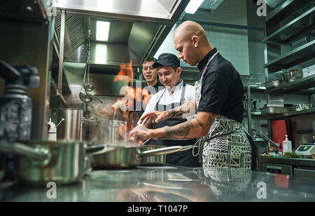 Extreme cooking. Profesional chef teaching his two young trainees how how to flambe food safely. Restaurant kitchen. Cooking process Stock Photo