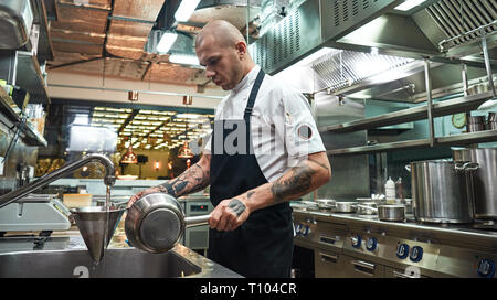 Cooling down. Serious professional chef holding cooked pasta in a colander under cold water in a restaurant kitchen. Cooking process Stock Photo