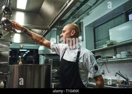Famous soup. SIde view of attractive bald chef in apron taking a ladle for soup cooking while standing in a restaurant kitchen. Cooking concept Stock Photo