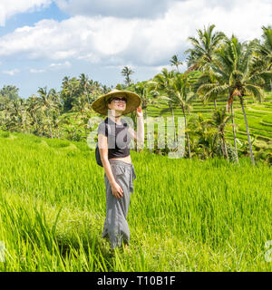 Relaxed fashionable caucasian female tourist wearing small backpack and traditional asian paddy hat walking among beautiful green rice fields and Stock Photo
