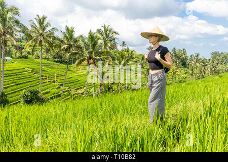 Relaxed fashionable caucasian female tourist wearing small backpack and traditional asian paddy hat walking among beautiful green rice fields and Stock Photo