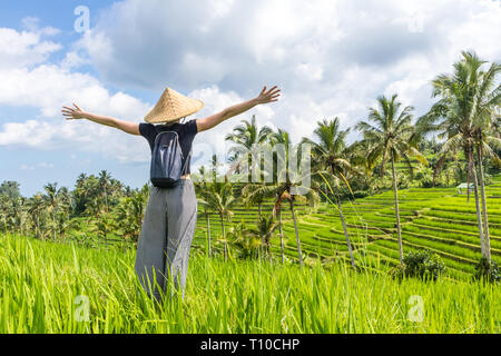 Relaxed fashionable female traveler wearing small backpack and traditional asian paddy hat, arms rised to sky, enjoying pure nature at beautiful green Stock Photo